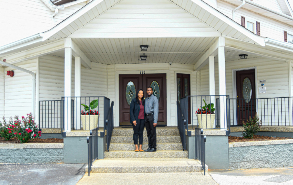 Two people standing side by side on the front steps of a funeral home.